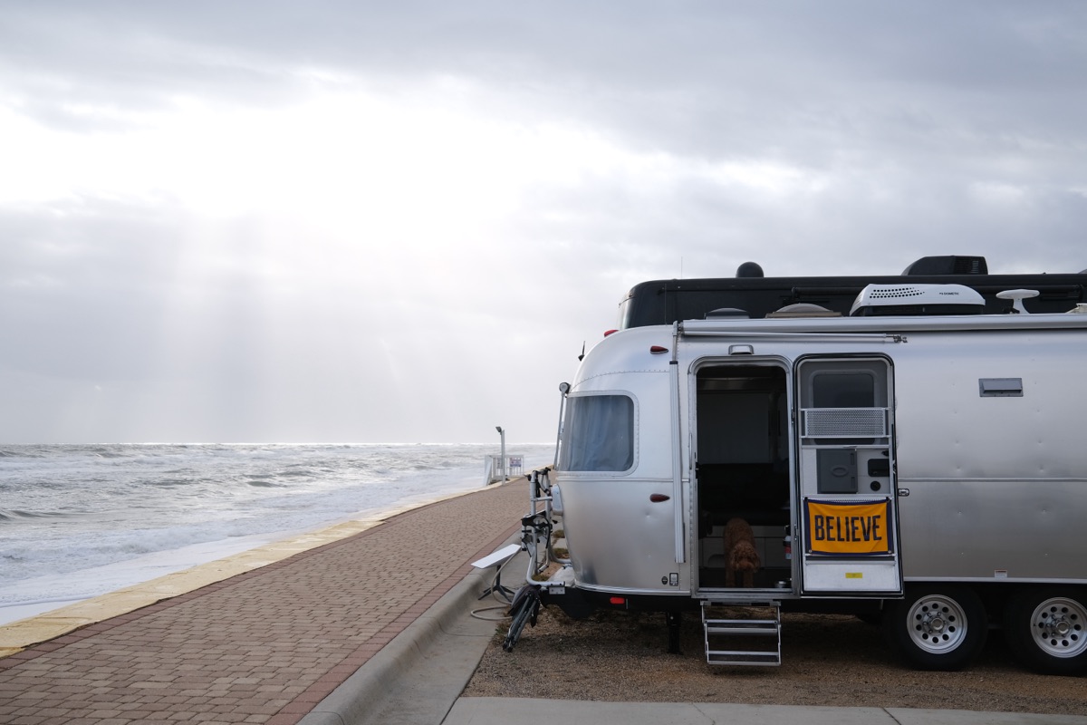 An Airstream trailer parked on a beach at dusk, ocean waves in the background and light shafts breaking through storm clouds overhead.