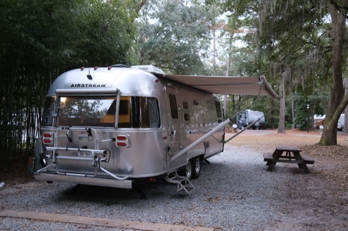 An Airstream trailer with its awning extended at a wooded campground site beneath Spanish-moss-draped trees, a picnic table alongside.