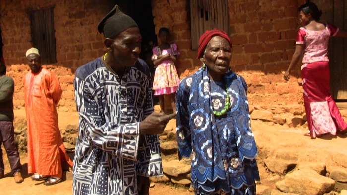 Two people in Ankara-print clothing talking in a terracotta-walled courtyard, a child visible nearby.