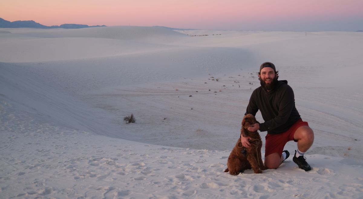 A bearded man in a dark hoodie kneeling on a white gypsum dune at dusk with one hand on an apricot curly-coated dog, a pink and lavender sunset band along the horizon behind them.